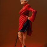 An older woman with silver hair stands tall in a bold red coat and black high heels. Her posture is fierce and unapologetic, one hand on her hip, the other holding a cane. She gazes to the side with sharp confidence, embodying strength, sass, and the power of aging boldly.