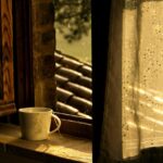 A white cup of tea resting on a sunlit windowsill, warm light filtering through rain-speckled glass, creating a quiet, contemplative scene.