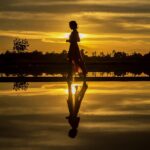 Silhouette of woman walking, her image reflected in the water, sun setting. Image by sasint on pixabay.