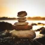 Stacked smooth river stones balanced in a small cairn at sunrise, warm golden light reflecting off the water behind them. Image by Randgruppe on Pixabay