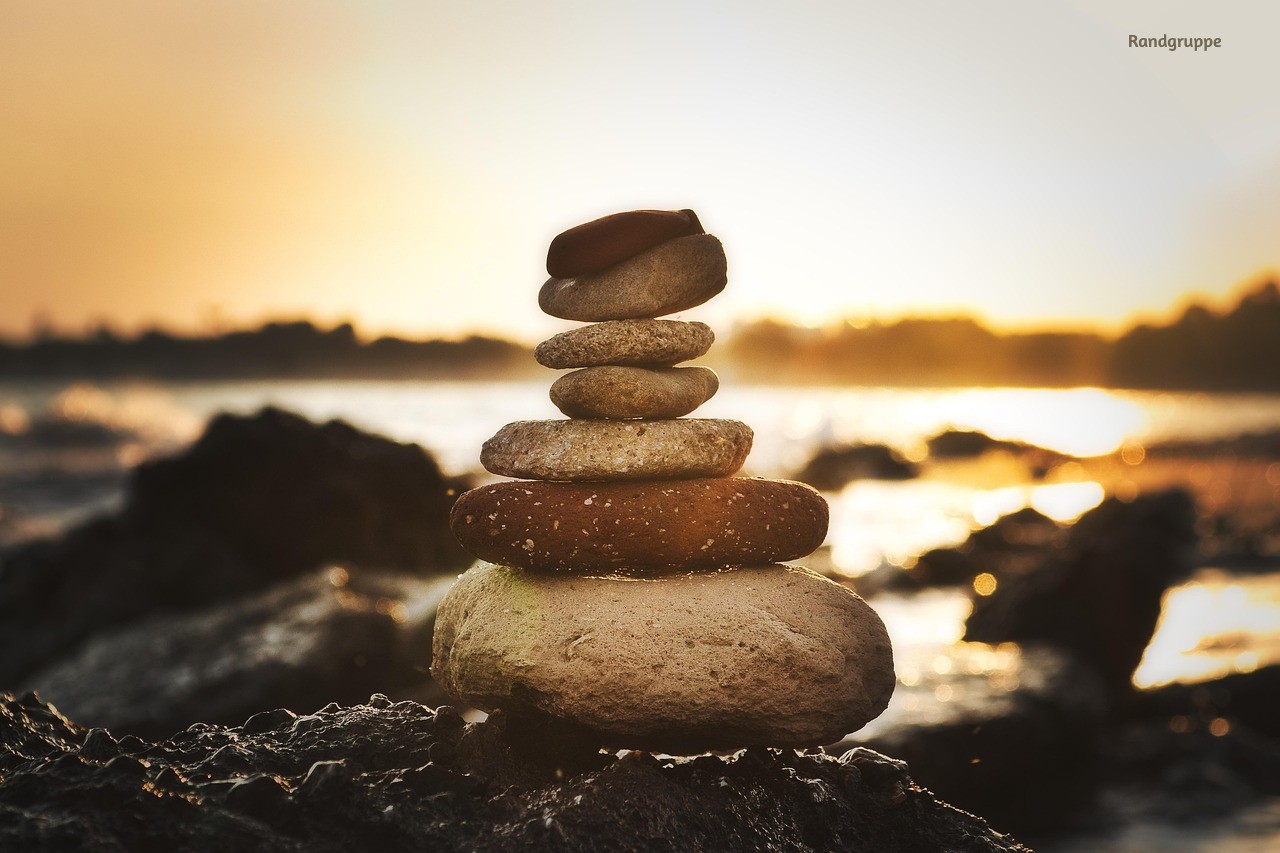 Stacked smooth river stones balanced in a small cairn at sunrise, warm golden light reflecting off the water behind them. Image by Randgruppe on Pixabay
