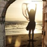 Woman standing in an open stone doorway, raising her arms toward the sunlight and sea beyond.