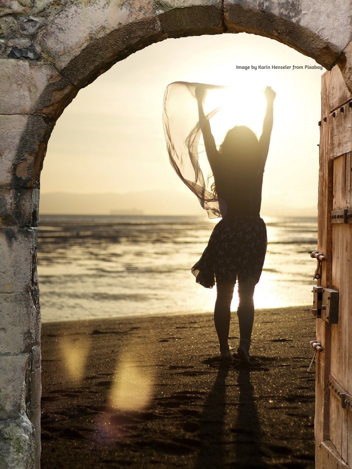 Woman standing in an open stone doorway, raising her arms toward the sunlight and sea beyond.